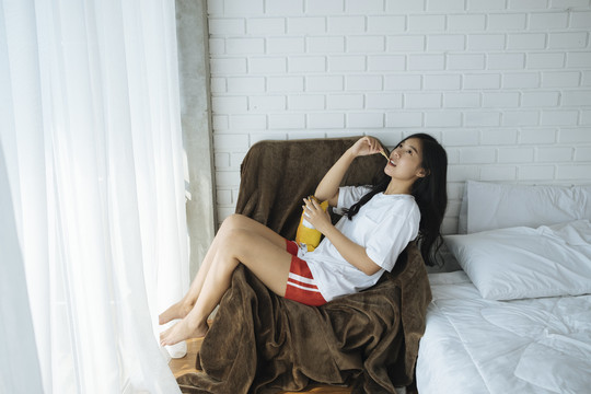 年轻的亚洲女士在舒适的沙发上吃薯片袋里的薯片。