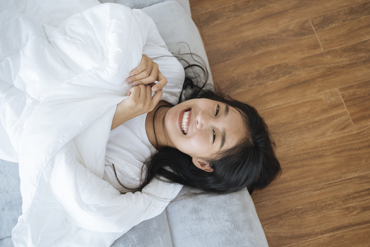 亚洲年轻女子躺在床上用白色毯子盖住身体的俯视图。快乐和微笑。