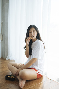年轻的亚洲女人坐在地板上，后面是白色的窗帘，吃着饼干盒里的饼干。