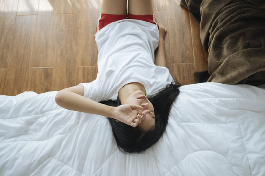 俯视图：年轻的亚洲妇女坐在地板上，躺在床沿上。睡错地方了。