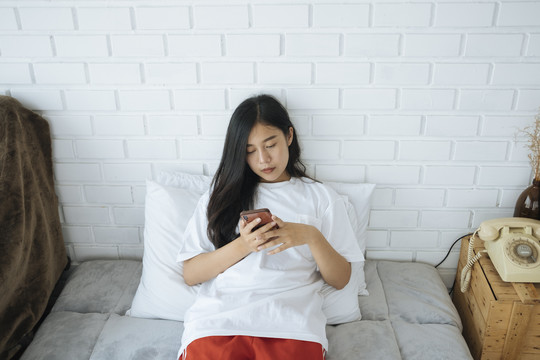 可爱的亚洲年轻女子躺在床上玩智能手机。和朋友聊天。