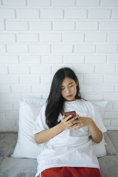 可爱的亚洲年轻女子躺在床上玩智能手机。和朋友聊天。