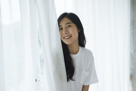 十几岁的泰国女孩站在卧室的白色窗帘后面。在家度假的概念。