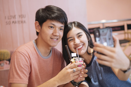 亚泰夫妇在甜点咖啡厅用智能手机自拍。男朋友拿着甜点冰淇淋。