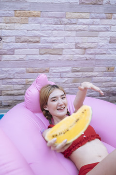 穿着红色比基尼的泰国美女躺在粉红色的泳衣上，手里拿着一块西瓜。