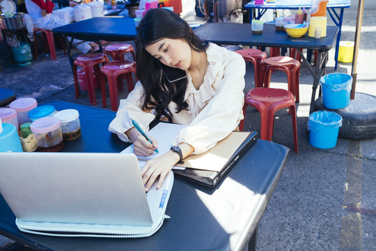 一位穿着休闲衬衫的泰国工作女商人在户外的街边小吃摊上工作。午餐时间工作。忙碌的。
