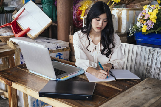 穿着休闲衬衫的泰国工作女商人在书上做笔记。在家里用笔记本电脑工作。