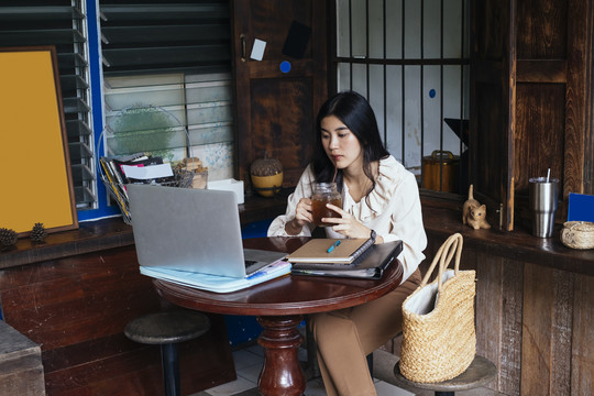 穿着休闲衬衫在家工作的泰国美女。用玻璃杯喝可乐和苏打水。