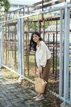 一位身穿白衬衫、背着书包的泰国年轻女子的肖像，斜靠在学校运动场附近的栅栏上。