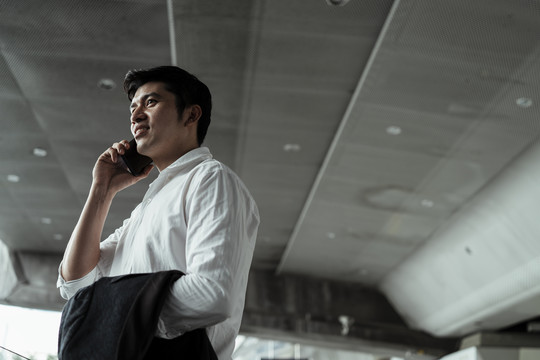 穿着白色正式衬衫的成熟亚洲商务人士在办公室外微笑着用智能手机交谈。