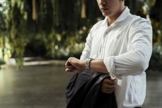 在花园里，一位穿着白色正式衬衫的成熟亚洲商人看着手腕上的手表。