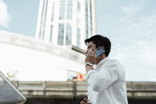 在大城市里，穿着白色正式衬衫的成熟亚洲商务人士用智能手机打电话。