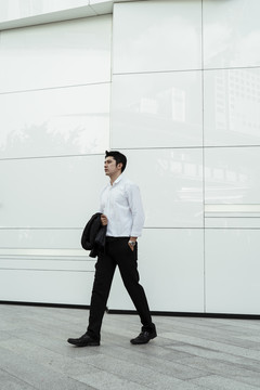 成熟的亚洲商人，穿着白色正式衬衫，手挽西服，走在白墙前。