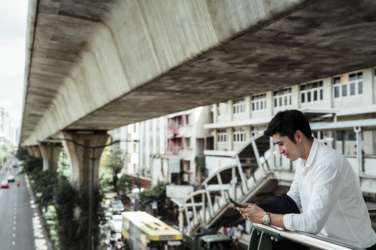 在曼谷市的天桥上，一个穿着白色正式衬衫的成熟的亚洲商人正在用智能手机微笑。