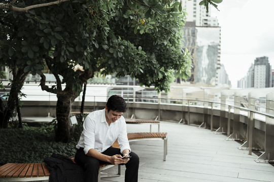 一个穿着白色正式衬衫的成熟的亚洲商人坐在屋顶的长凳上，在大树下看着城市玩着智能手机，微笑着。