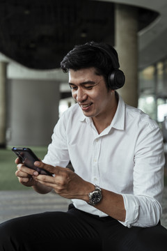 穿着白色正式衬衫，戴着耳机，从智能手机播放音乐的亚洲成熟商务人士特写镜头。用酷炫的小玩意欣赏这首歌。