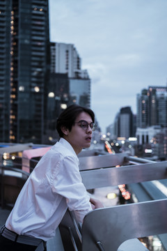 身穿白衬衫、戴着眼镜的黑发男子站在城市楼顶，傍晚时分欣赏和感受城市的风和夜景。