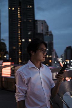 一个穿着白衬衫戴着眼镜的黑发男人站在屋顶上，用他的智能手机设置了有线路障，背景是城市里的建筑、道路、