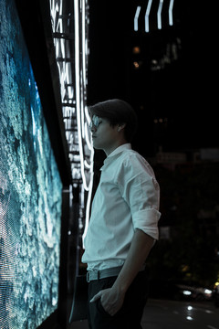 晚上，身穿白衬衫、戴眼镜的黑发男子站在市区商场前的LED显示屏前。