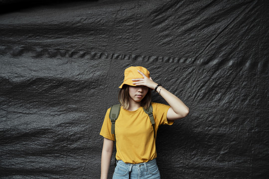 身穿深黄色t恤的亚洲女孩站在黑色背景前，用深黄色帽子遮住脸。