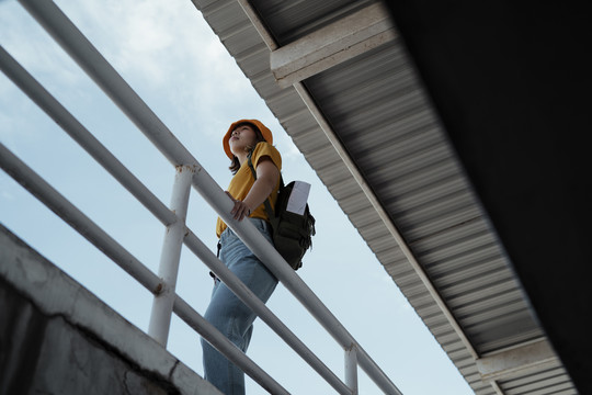以屋顶和天空为背景，站在天桥上的黄色背包女子背着绿色背包的低角度镜头。