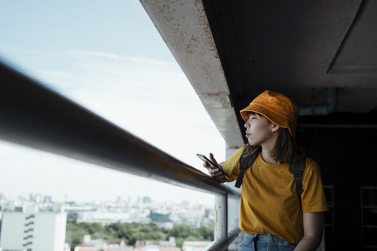 黄色背包女，背着绿色背包，坐在高楼的停车场上，以扶手为主线，眺望着城市景色。