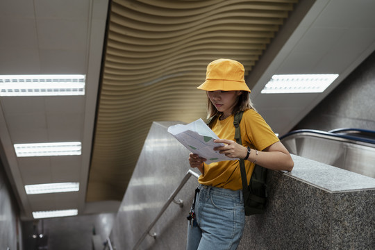 黄色背包女背着绿色背包靠在通往地铁站的楼梯扶手上拿着地图。