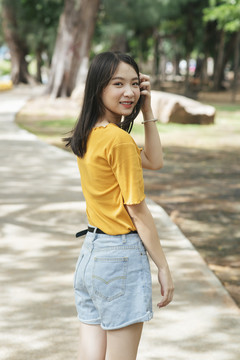 夏天，穿着黄色t恤的黑色长发女孩站在公园的小路上。
