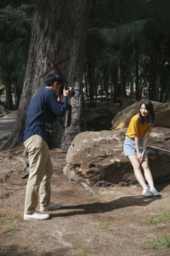 男摄影师拍了一张十几岁的女模特坐在公园大树旁的岩石上的照片。