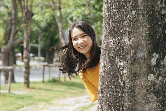 活泼的黑发女孩站在树后对着镜头微笑。