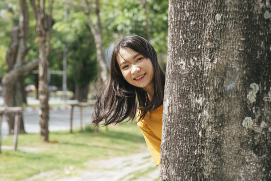 活泼的黑发女孩站在树后对着镜头微笑。