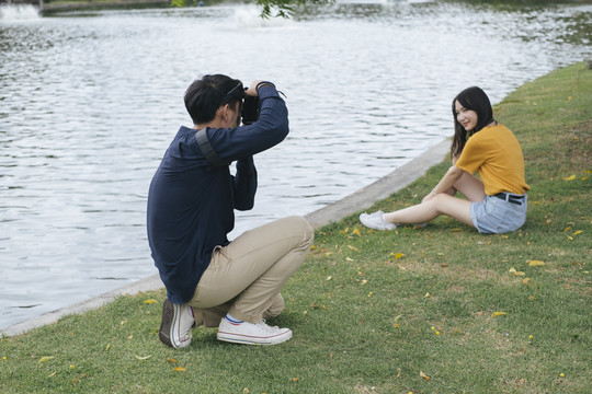 男摄影师坐在公园里的湖边给十几岁的模特拍照。