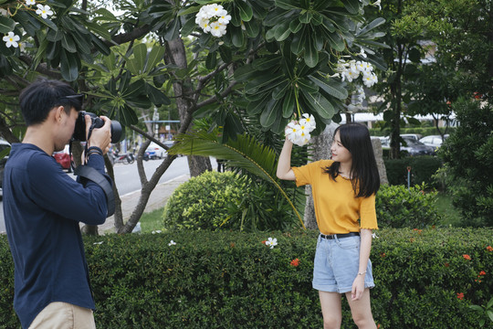 男摄影师拿着鲜花和相机，在鸡蛋花树旁为十几岁的模特女孩拍照。
