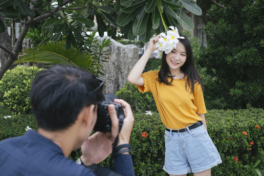 男摄影师拿着鲜花和相机，在鸡蛋花树旁为十几岁的模特女孩拍照。