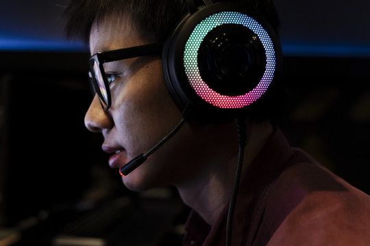 特写，侧视，头像的书呆子年轻亚洲人戴着眼镜和耳机或耳机，在电脑上玩网吧，暗室。