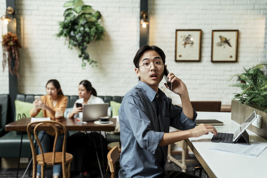 一个年轻的亚洲男人正在打电话，惊讶地看着相机，正在使用平板电脑。
