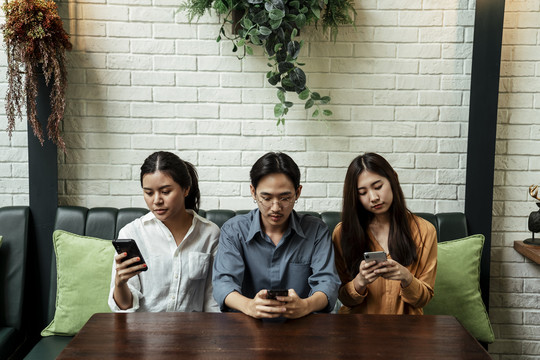三个年轻的亚洲人坐在沙发上玩智能手机，无视坐在他们旁边的朋友。社交成瘾。