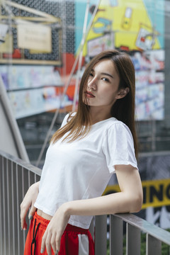 时尚写真-美丽的亚洲年轻女子，穿着白衬衫和红色运动裤，斜靠在天桥扶手上。