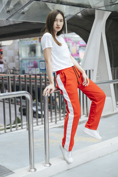 时尚写真-美丽的亚洲年轻女子穿着白衬衫和红色运动裤坐在扶手天桥上。