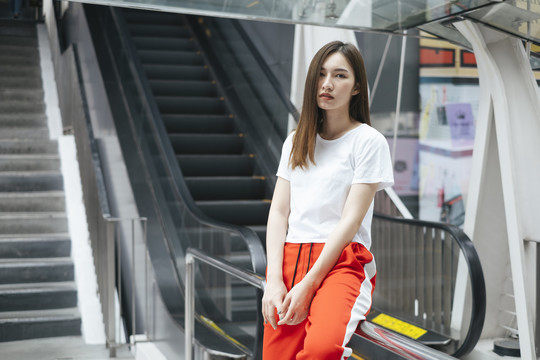 时尚写真-穿着白衬衫和红色运动裤的美丽亚洲年轻女子在自动扶梯前摆姿势。