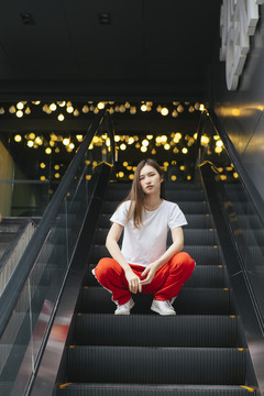 时尚写真——坐在自动扶梯上的穿着白衬衫和红色运动裤的美丽亚洲年轻女子。后面有漂亮的波基背景。