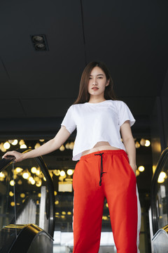 时尚写真——站在自动扶梯上的穿着白衬衫和红色运动裤的美丽亚洲年轻女子。背景是美丽的波基。