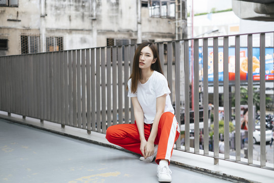 时尚写真-美丽的亚洲年轻女子穿着白衬衫和红色运动裤坐在城市的天桥上。