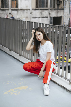 时尚写真-美丽的亚洲年轻女子穿着白衬衫和红色运动裤坐在城市的天桥上。