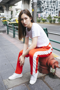 时尚写真——穿着白衬衫和红色运动裤的亚洲美女坐在人行道上的红色灭火水管上。