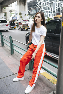 时尚写真-穿着白衬衫和红色运动裤坐在人行道扶手上的美丽亚洲年轻女子。