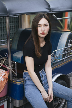 时尚写真-美丽的时髦的亚洲年轻女子在黑色衬衫和牛仔裤裤子坐在图克图克座位在曼谷。