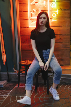 时尚写真-穿着黑色衬衫和牛仔裤的亚洲时髦年轻女子坐在霓虹灯背景下的椅子上。