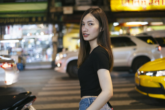 时尚写真-美丽的时髦亚洲年轻女子在黑色衬衫和牛仔裤裤子走在马路对面的夜晚在曼谷。