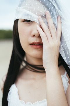 特写-年轻的亚洲长黑发女子新娘的肖像，戴着白色面纱，在户外用手遮住眼睛。